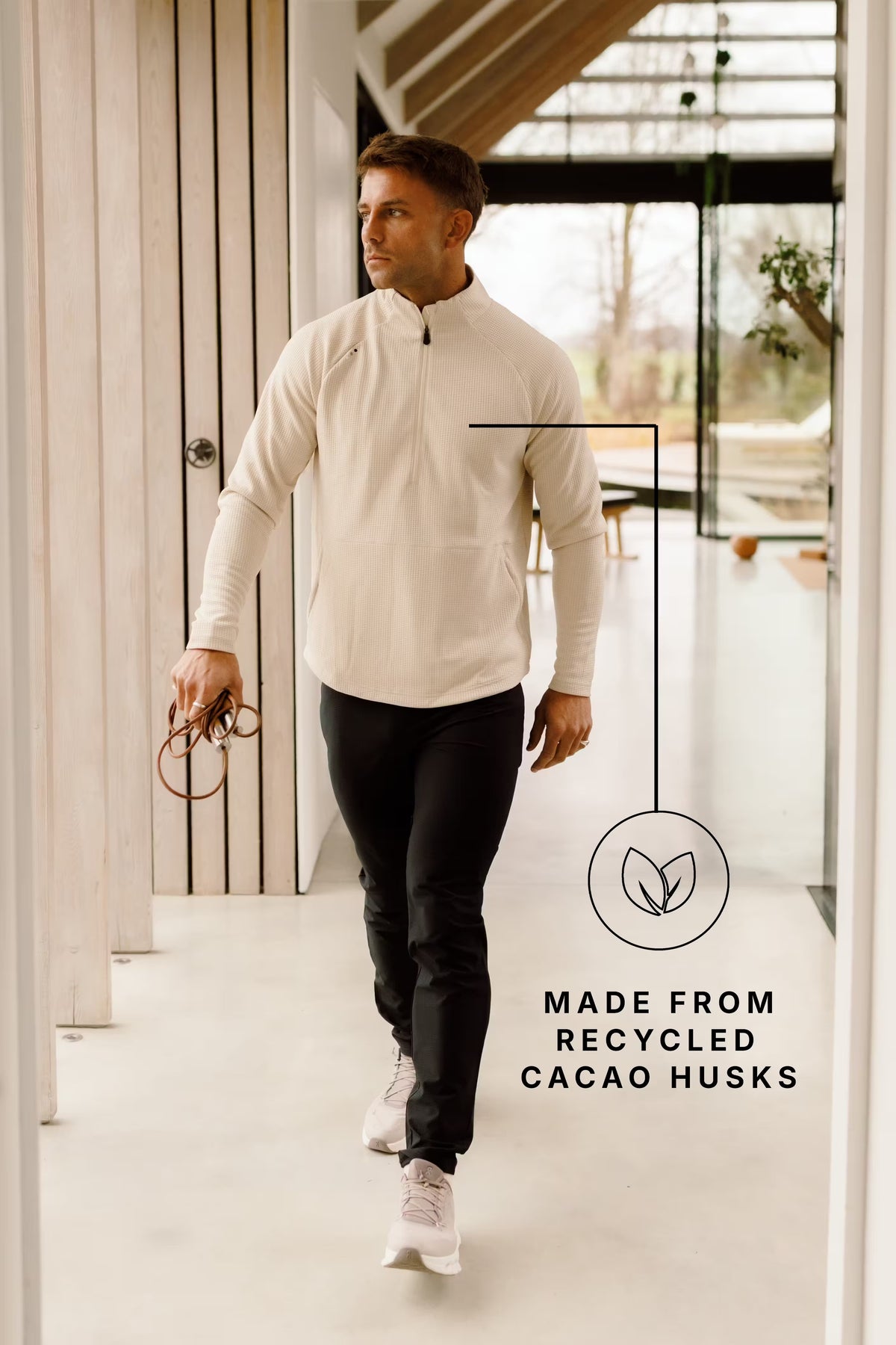 Man in a cream PRO | ORA half zip and black trousers walking through a bright, timber-lined hallway