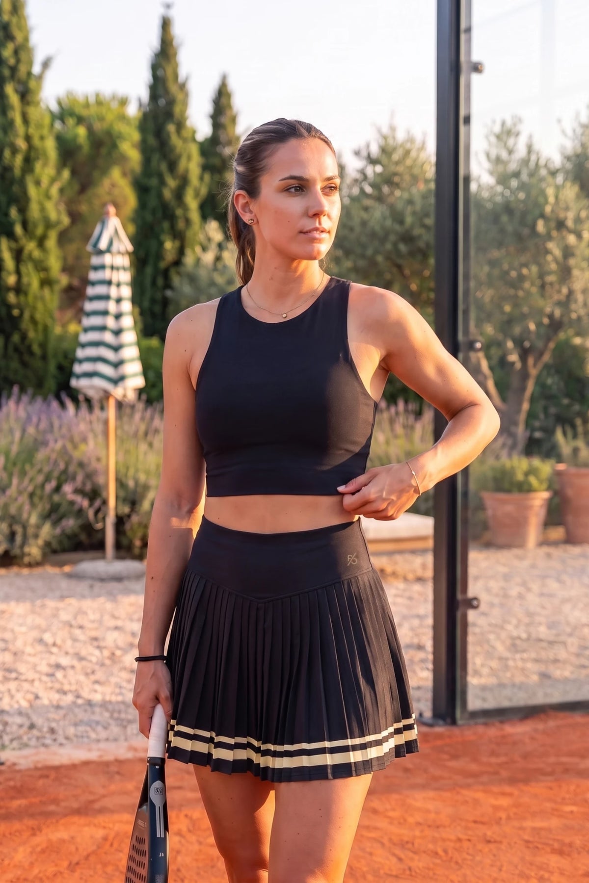 The Aura Court Skirt in Black Onyx worn on a padel court, model standing next to a striped parasol #Color_Black Onyx